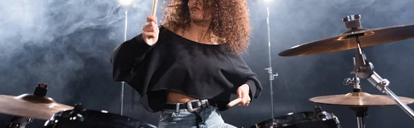 Cropped view of curly woman with drumsticks playing on drum kit with smoke on background, banner — Stock Photo