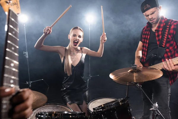 KYIV, UKRAINE - AGOSTO 25, 2020: Mulher loira com baquetas gritando enquanto se senta no kit de bateria perto do guitarrista com guitarra enevoada e embaçada em primeiro plano — Fotografia de Stock