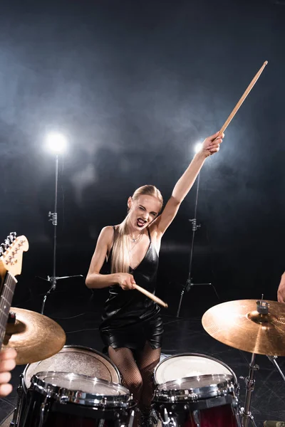Blonde woman with outstretched hand winking while sitting near drum kit with backlit on blurred foreground — Stock Photo