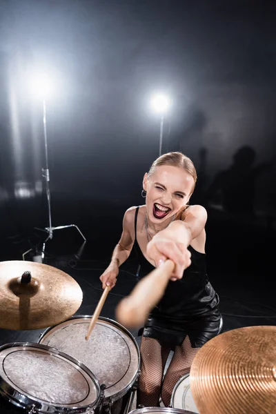 Hochwinkelaufnahme der aufgeregten Blondine, die mit Drumstick zeigt, während sie am Schlagzeug sitzt, mit Hintergrundbeleuchtung auf verschwommenem Vordergrund — Stockfoto