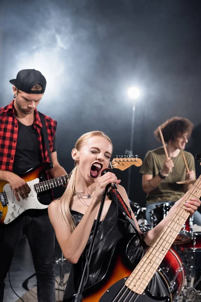 KYIV, UKRAINE - 25 de agosto de 2020: Vocalista femenina de la banda de rock gritando en el micrófono cerca del guitarrista con el batería retroiluminado y borroso en el fondo - foto de stock