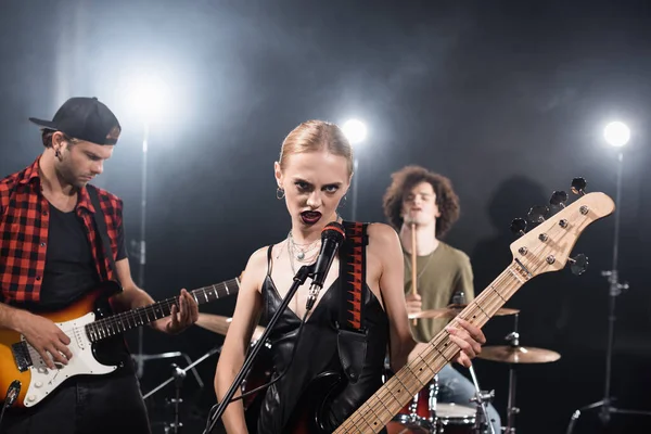 KYIV, UCRANIA - 25 de agosto de 2020: Mujer enojada con la guitarra eléctrica mirando a la cámara cerca del guitarrista con el batería retroiluminado y borroso en el fondo - foto de stock