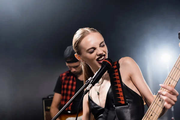 KYIV, UKRAINE - AUGUST 25, 2020: Rock band vocalist with electric guitar singing in microphone with backlit and blurred man on background — Stock Photo