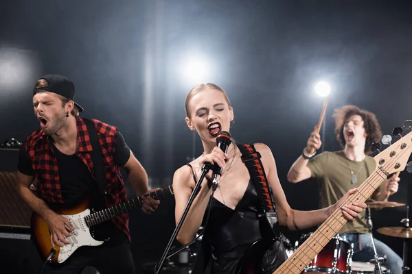 KYIV, UKRAINE - AUGUST 25, 2020: Blonde vocalist with electric guitar singing in microphone near guitarist and drummer with backlit on blurred background — Stock Photo
