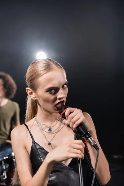 Angry blonde woman singing while touching microphone rack with backlit and blurred drummer on background — Stock Photo