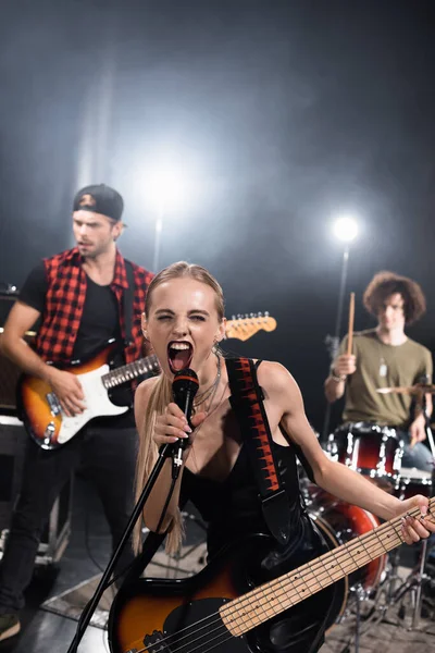 КИЕВ, УКРАИНА - 25 августа 2020 года: Женщина-вокалистка рок-группы с электрогитарой кричит в микрофоне с подсветкой и размытыми музыкантами на заднем плане — стоковое фото