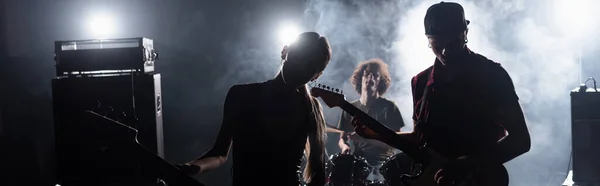 KYIV, UKRAINE - AUGUST 25, 2020: Rock band guitarists standing near combo amplifiers and drummer sitting at drum kit with backlit and smoke on background, banner — Stock Photo