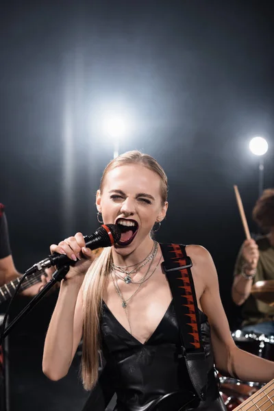 Blonde woman with bass guitar screaming in microphone with backlit and blurred musicians on background — Stock Photo