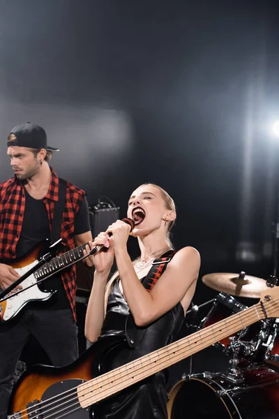 KYIV, UCRANIA - 25 de agosto de 2020: Mujer rubia con guitarra eléctrica, inclinada hacia atrás mientras grita en el micrófono cerca del guitarrista y el kit de batería con retroiluminación en el fondo - foto de stock