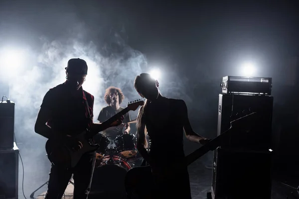 KYIV, UKRAINE - AUGUST 25, 2020: Rock band guitarists standing near combo amplifiers and drummer sitting at drum kit with backlit and smoke on background — Stock Photo