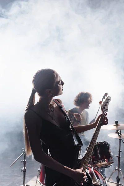 KYIV, UKRAINE - AUGUST 25, 2020: Woman playing electric guitar with smoke and blurred drummer with drum kit on background — Stock Photo
