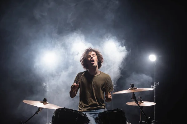 Emocionado baterista rizado con la boca abierta sentado en el kit de batería con humo y retroiluminado en negro - foto de stock