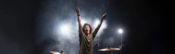 Musicien bouclé excité avec les mains dans l'air assis près des cymbales avec de la fumée et rétroéclairé sur le fond, bannière — Photo de stock