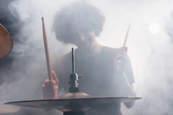 Smiling curly drummer with drumsticks playing drums in smoke — Stock Photo