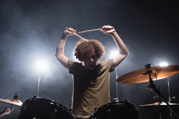 Músico encaracolado gritando, enquanto segurando baquetas e sentado no kit de bateria com retroiluminação no fundo — Fotografia de Stock