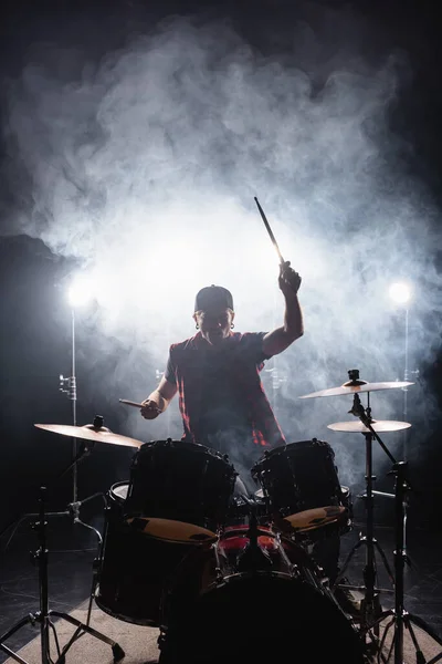 Homem tocando bateria enquanto sentado no kit de bateria com retroiluminação e fumaça no fundo — Fotografia de Stock