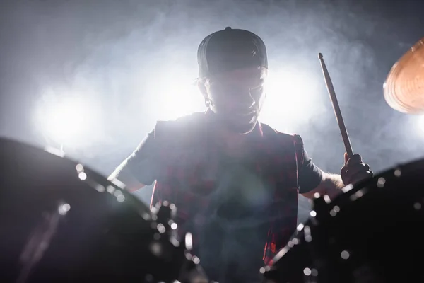 Músico de banda de rock con baqueta tocando la batería con humo y retroiluminado en el fondo - foto de stock