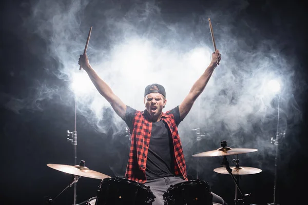 Shouting drummer with hands in air sitting at drum kit with backlit and smoke on black — Stock Photo
