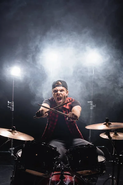 Кричащий барабанщик со скрещенными барабанными палочками смотрит в камеру, сидя за барабанной установкой с дымом и подсветкой на заднем плане — стоковое фото