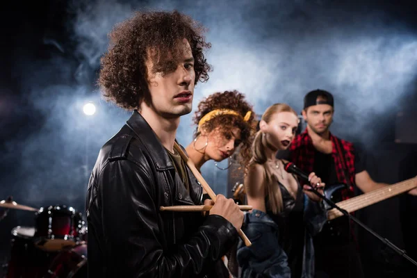 KYIV, UKRAINE - AUGUST 25, 2020: Curly musician with drumsticks looking at camera with smoke and blurred members of rock band on background — Stock Photo