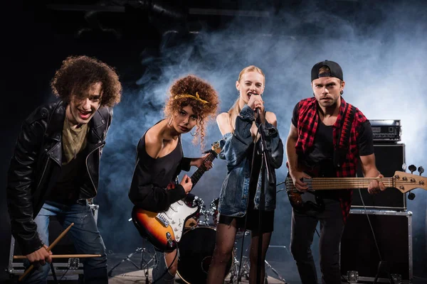 KYIV, UKRAINE - AUGUST 25, 2020: Female vocalist singing, while standing near rock band members with bass guitars and drumsticks, with smoke on black — Stock Photo