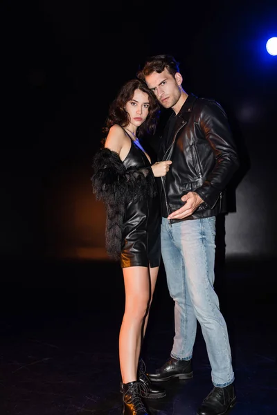 Curly brunette woman touching boyfriend jacket, while looking at camera with backlit on black — Stock Photo