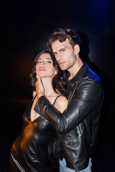 Confident man in leather jacket touching girlfriend neck, while looking at camera on black — Stock Photo