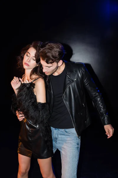 Man in leather jacket embracing sexy girlfriend in dress on black — Stock Photo