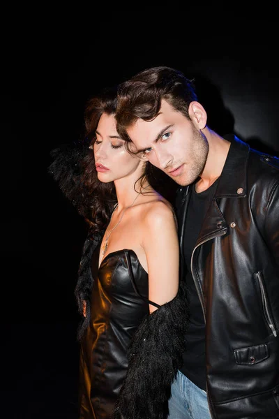 Seductive woman with naked shoulder standing near man in leather jacket looking at camera on black — Stock Photo