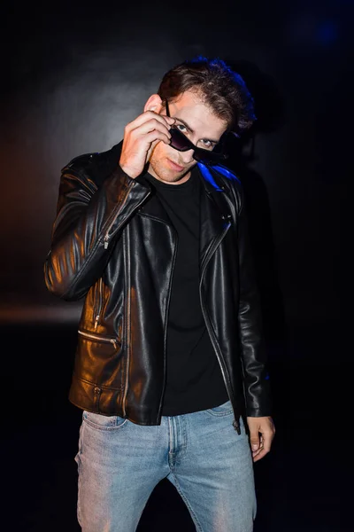 Front view of sexy man holding eyeglasses frame, while wearing leather jacket on black — Stock Photo