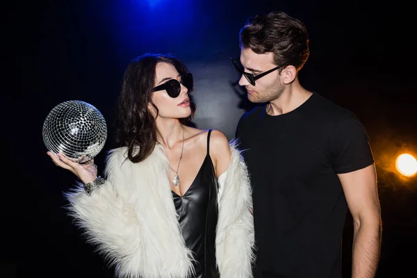 Sexy brunette woman with disco ball standing near man in eyeglasses with backlight on black — Stock Photo