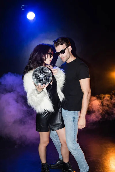 Femme sexy avec boule disco debout près de l'homme dans des lunettes avec de la fumée et rétro-éclairé sur noir — Photo de stock