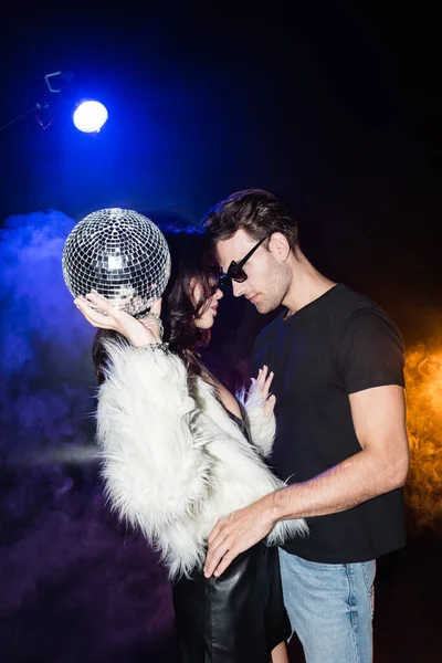 Donna bruna con palla da discoteca in aria toccare camicia fidanzato con fumo e retroilluminato su nero — Foto stock