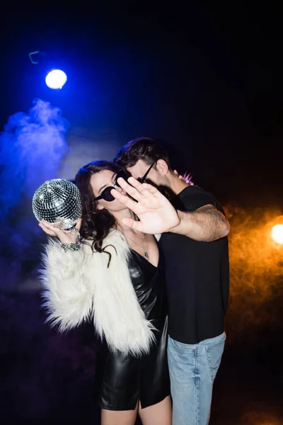 Hombre cerrando la cámara con la mano, mientras abraza a la mujer sosteniendo la bola disco con retroiluminación y humo en negro - foto de stock