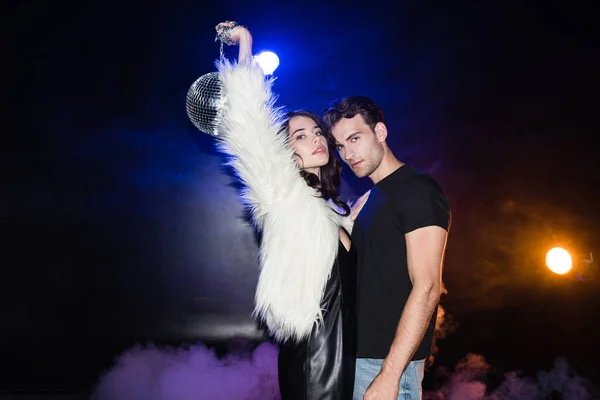 Brunette woman holding disco ball in air, while embracing with boyfriend and looking at camera with smoke and backlit on black — Stock Photo