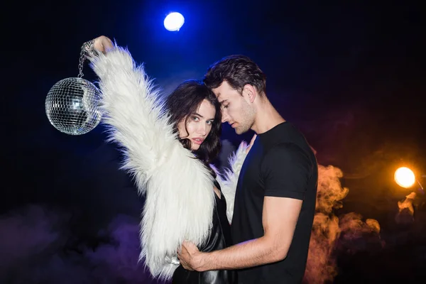 Leidenschaftlicher Mann umarmt Frau, hält Discokugel in der Luft und blickt in Kamera mit Rauch und Hintergrundbeleuchtung auf schwarz — Stockfoto