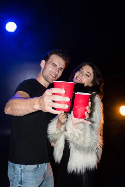 Casal feliz segurando copos de plástico vermelho e olhando para a câmera com retroiluminado em preto, no fundo borrado — Fotografia de Stock
