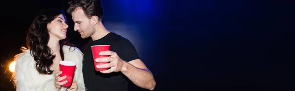 Passionate man standing near seductive woman, looking at lips, while holding plastic cup with backlit on black, banner — Stock Photo