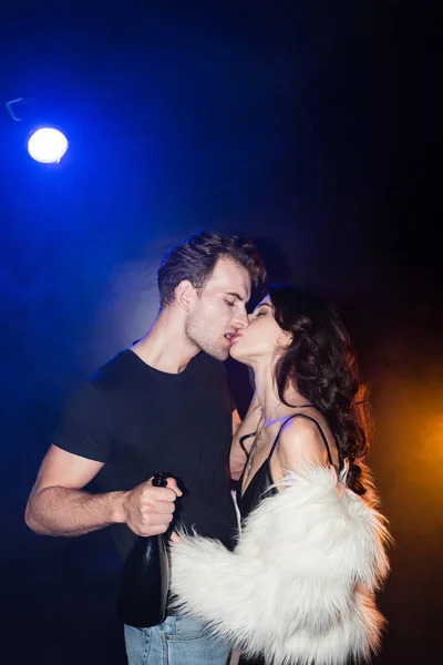 Mulher sedutora beijando namorado com garrafa de champanhe com retroiluminado em preto — Fotografia de Stock