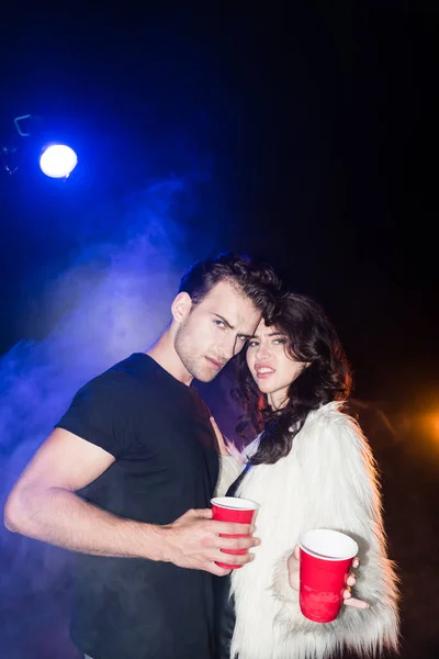 Irritated young adult couple looking at camera, holding plastic cups, while embracing with smoke and backlit on black — Stock Photo