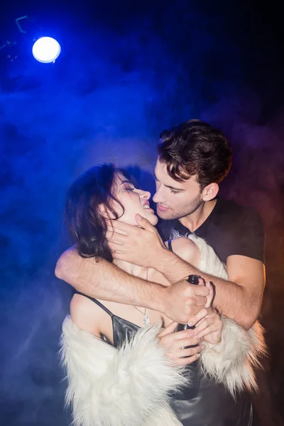 Passionate man with champagne bottle embracing and looking at lips of sexy woman with smoke and backlit on black — Stock Photo