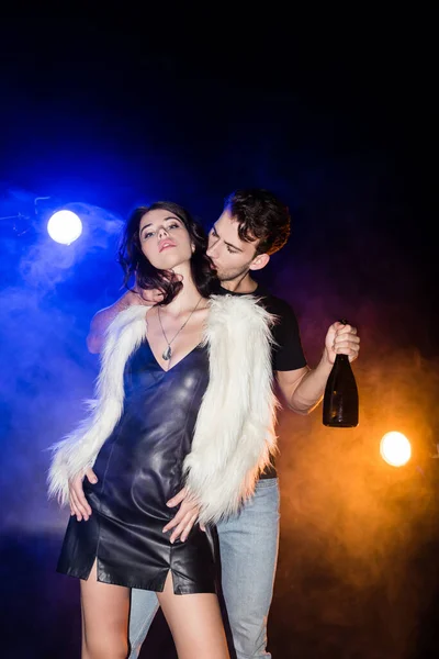 Low angle view of passionate man with champagne bottle touching woman neck with smoke and backlit on black — Stock Photo