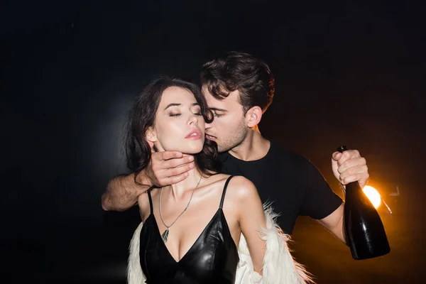 Страстный мужчина с бутылкой шампанского трогает сексуальную женщину за шею с подсветкой на черном — стоковое фото