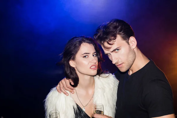 Irritated couple with glasses of champagne hugging and looking at camera with backlit on black — Stock Photo