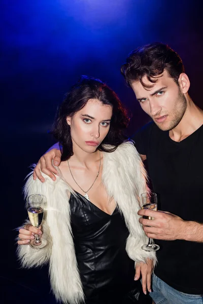 Ernsthafter Mann mit Glas Champagner umarmt Freundin in Kleid und weißer Jacke mit Hintergrundbeleuchtung auf Schwarz — Stockfoto