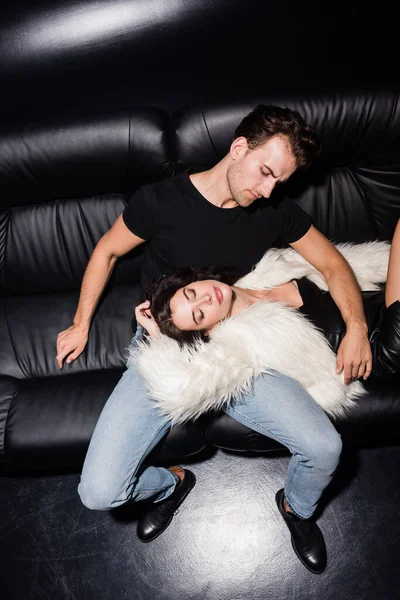 Vista de ángulo alto de la mujer sonriente con la mano detrás de la cabeza acostado en el hombre en ropa casual en el sofá en el club nocturno - foto de stock