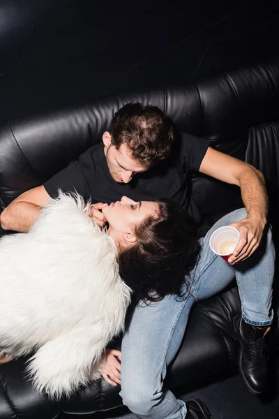 Vue grand angle de l'homme avec le pied sur le canapé, toucher le menton de la femme, tout en tenant une tasse de café en plastique dans la boîte de nuit — Photo de stock