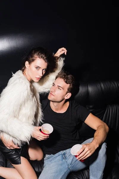 Selbstbewusster Mann umarmt brünette Frau, die auf Knien auf Sofa sitzt und in Nachtclub in die Kamera schaut — Stockfoto
