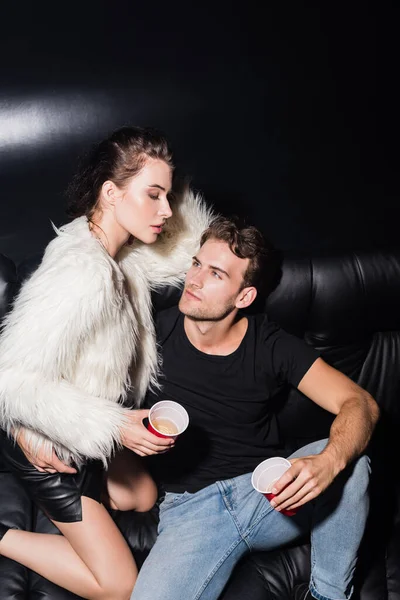 Selbstbewusster Mann umarmt verführerische Frau, sitzt auf Sofa und hält Plastikbecher Kaffee in Nachtclub — Stockfoto