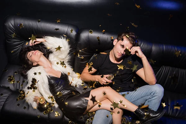 Giovane uomo adulto guardando la macchina fotografica, seduto sul divano vicino alla donna sdraiato con un bicchiere di champagne, mentre i coriandoli cadono in discoteca — Foto stock
